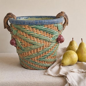 May include: A ceramic basket-style planter with a woven design in shades of green and tan. The planter features decorative handles and is adorned with sculpted fruit and leaves. Three pears rest on a linen cloth nearby.