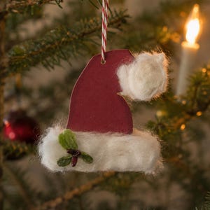 May include: A flat, dark red Santa hat ornament featuring a white fluffy trim and a matching white pom-pom. The hat is accented with a small green leaf and dark berry detail on the brim and hangs from a red and white twisted string.