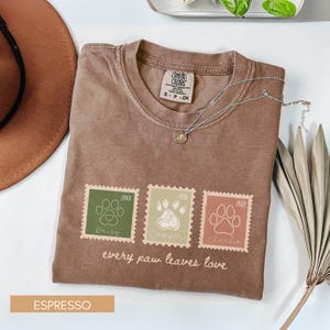 May include: An espresso-colored t-shirt featuring three rectangular stamp-style graphics with paw prints, names, and years. The text reads "every paw leaves love" below the stamps. The shirt has a Comfort Colors label at the inner neckline.