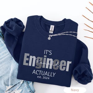 Peut inclure: Un sweat-shirt bleu marine à col rond avec le texte blanc et gris IT'S Engineer ACTUALLY est. 2024, incluant le prénom Ronald écrit en lettres cursives sur le mot Engineer. Le vêtement possède des manches longues et un col côtelé.