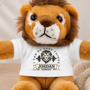 May include: A plush lion toy with golden-brown fur and a dark brown mane, wearing a white t-shirt. The shirt features a graphic of a lion head with a barbell and the text I CAN DO HARD THINGS, JORDAN, and FEARLESS printed in black ink.