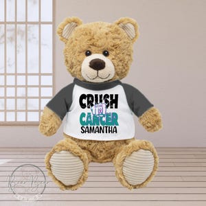 May include: A brown teddy bear wearing a white and grey shirt with the text "CRUSH CANCER" and the name "SAMANTHA" printed on it.