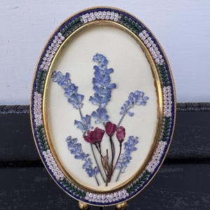 May include: Oval-shaped pressed flower art in a gold-toned frame. The frame has a blue and white border. The artwork features blue and purple pressed flowers on a cream-colored background. The frame is supported by small gold-toned feet.