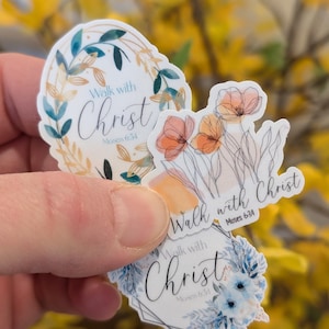 Pode incluir: Três autocolantes recortados com desenhos florais e o texto Walk with Christ Moses 6:34. Um autocolante tem uma coroa de folhas douradas e azul-petróleo, um tem papoilas de linhas cor de laranja e um tem um arranjo floral azul.