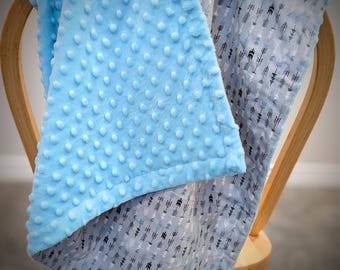 Puede incluir: Una manta para bebé sobre una silla de madera. La capa superior es de tela azul claro texturizada con puntos en relieve. La parte inferior presenta una tela gris con un patrón de flechas negras y azules.