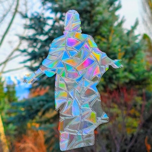 May include: A holographic figure of a person with outstretched arms, resembling a religious icon. The figure is composed of iridescent, geometric shapes that reflect a spectrum of colors. The background features blurred green foliage and a blue sky.