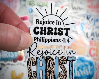 May include: A die-cut vinyl sticker featuring the text Rejoice in CHRIST Philippians 4:4 in black and blue with a leopard print pattern on the word CHRIST, topped with a black sunburst graphic, set against a background of similar stickers.