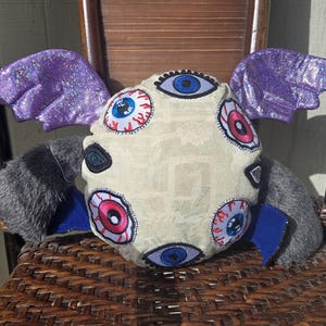 May include: A whimsical plush creature with a cream-colored body adorned with multiple eye appliqués. It features purple glitter wings, gray fur arms, and blue accents. The plush is seated on a woven chair.