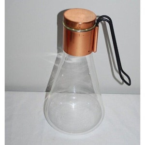 May include: A clear glass pitcher with a copper lid and a black metal handle. The pitcher has a rounded bottom and a wide mouth.