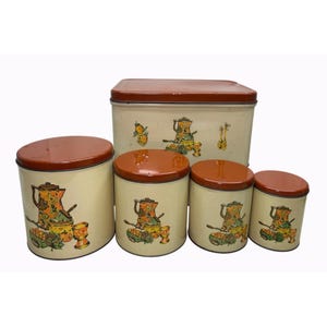 May include: A set of five cream-colored metal storage canisters with rust-colored lids. The containers feature a repeating graphic illustration of a coffee pot, a bowl, and various fruits in shades of orange, green, and brown on the exterior surface.