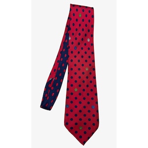 May include: A red necktie featuring a pattern of dark blue polka dots and small, scattered circular emblems in various colors including green, blue, and white. The tie is folded over itself, revealing a dark blue underside with red polka dots.