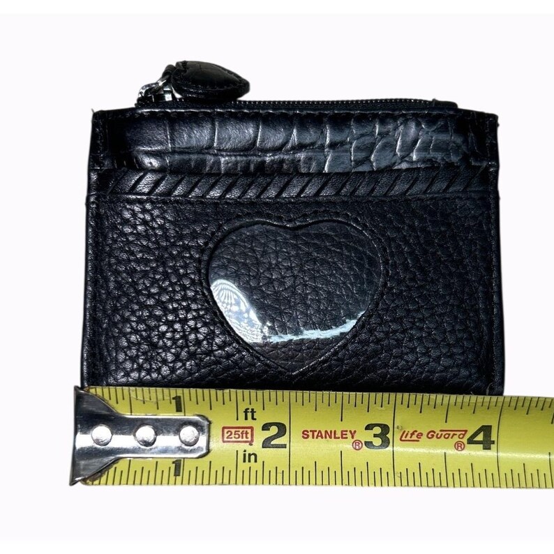 Puede incluir: Tarjetero de cuero negro con un recorte en forma de coraz&oacute;n y una cremallera en la parte superior. El tarjetero mide aproximadamente 10 cm de ancho. El dise&ntilde;o incluye un acabado de cuero texturizado y un ribete decorativo.
