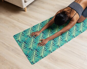 Könnte beinhalten: Eine grüne und blaue Yogamatte mit einem sich wiederholenden Muster aus tropischen Blättern. Die Matte ist rechteckig und liegt auf einem hellen Holzboden. Die Matte wird zum Dehnen verwendet.