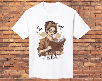 Könnte beinhalten: Weißes T-Shirt mit einer braunen Illustration einer Frau, die ein Buch liest, eine Brille und eine Schleife trägt. Der Text "In my bookish era" befindet sich über und unter der Illustration. Das T-Shirt wird vor einer Backsteinmauer präsentiert.