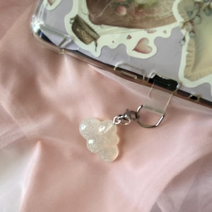 May include: A clear phone case with a floral design and a small, sparkly, cloud-shaped charm. The charm is attached to the case with a silver-colored clasp. The background is a soft, pink fabric.