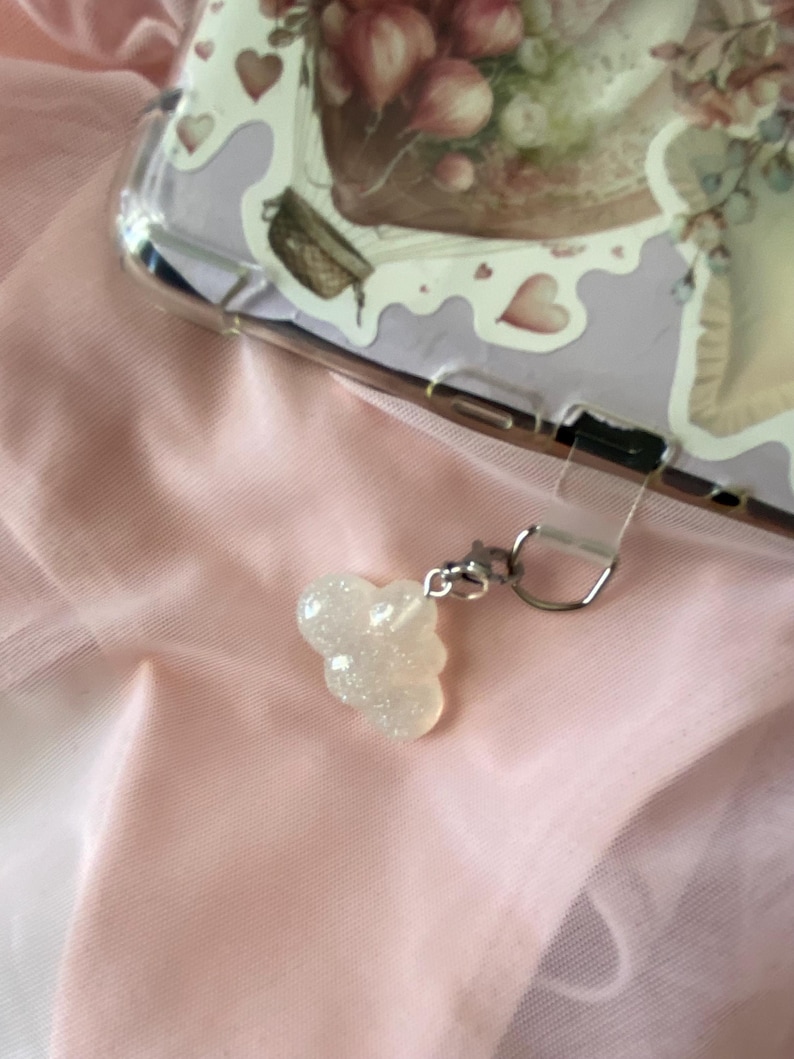 May include: A clear phone case with a floral design and a small, sparkly, cloud-shaped charm. The charm is attached to the case with a silver-colored clasp. The background is a soft, pink fabric.