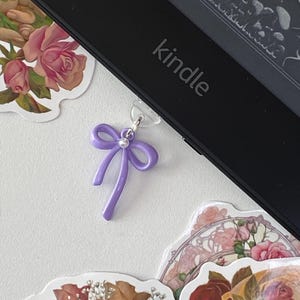 May include: A purple bow bookmark with a pearl accent attached to a Kindle e-reader.