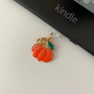 May include: A gold-colored charm in the shape of a red pumpkin with a green leaf, attached to a clear plastic piece. The charm is next to a black Kindle device with the word "kindle" printed on it.