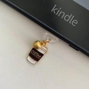 May include: A gold-colored coffee cup charm with the word "coffee" in black lettering, attached to a clear plastic piece. The charm is next to a black Kindle device with the word "kindle" printed on it.