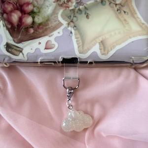May include: A clear phone case with a decorative sticker featuring floral and pillow designs. A silver-colored charm with a glittery cloud pendant is attached to the case. The background is a pink fabric.