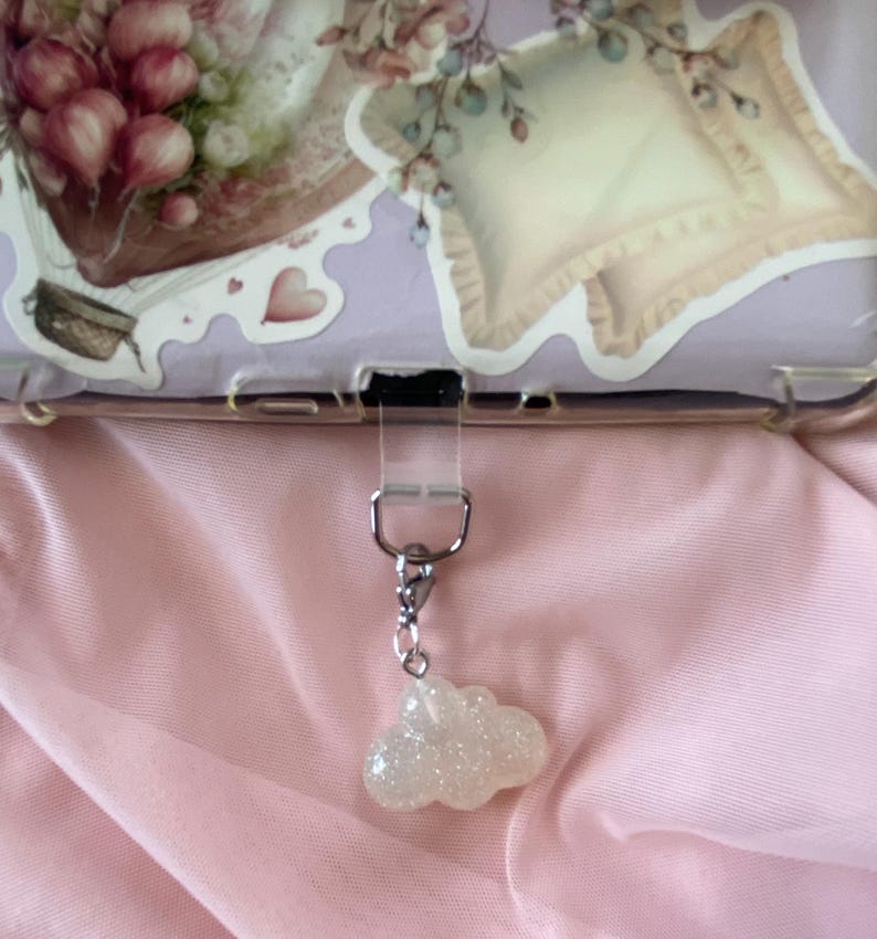 May include: A clear phone case with a decorative sticker featuring floral and pillow illustrations. A silver-toned charm with a glittery cloud pendant hangs from the case. The background is a pink fabric.