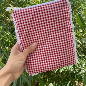 May include: A red and white gingham pattern quilted pouch with a white lace trim. The pouch has a zipper closure and a diamond pattern. The background is a lush green with white flowers.