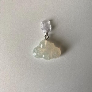 May include: A cloud-shaped earring with a clear plastic clip. The cloud is a light, glittery color, and is attached to the clip with a small silver-colored ring. The earring is set against a white background.