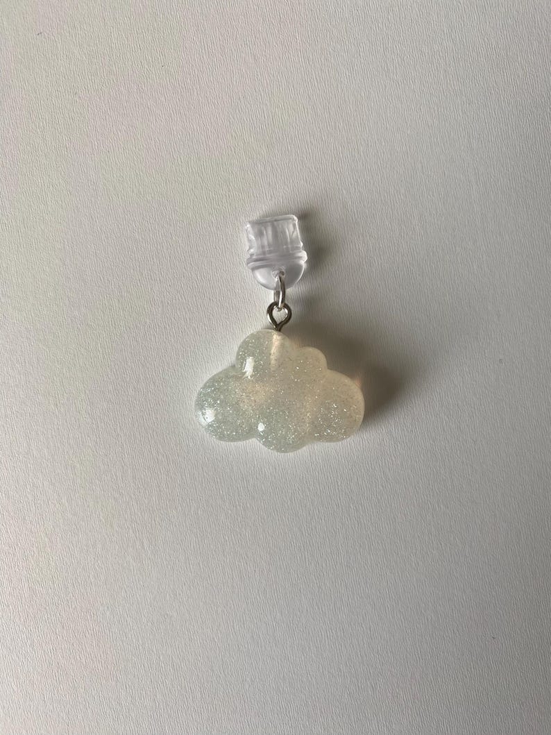 May include: A cloud-shaped earring with a clear plastic clip. The cloud is a light, glittery color, and is attached to the clip with a small silver-colored ring. The earring is set against a white background.