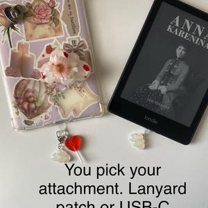 May include: A clear phone case decorated with stickers, including a rose, envelope, and candles. A Kindle e-reader displays the cover of "Anna Karenina." Charms include a red heart lollipop and a cloud. Text reads "You pick your attachment. Lanyard patch or USB-C."