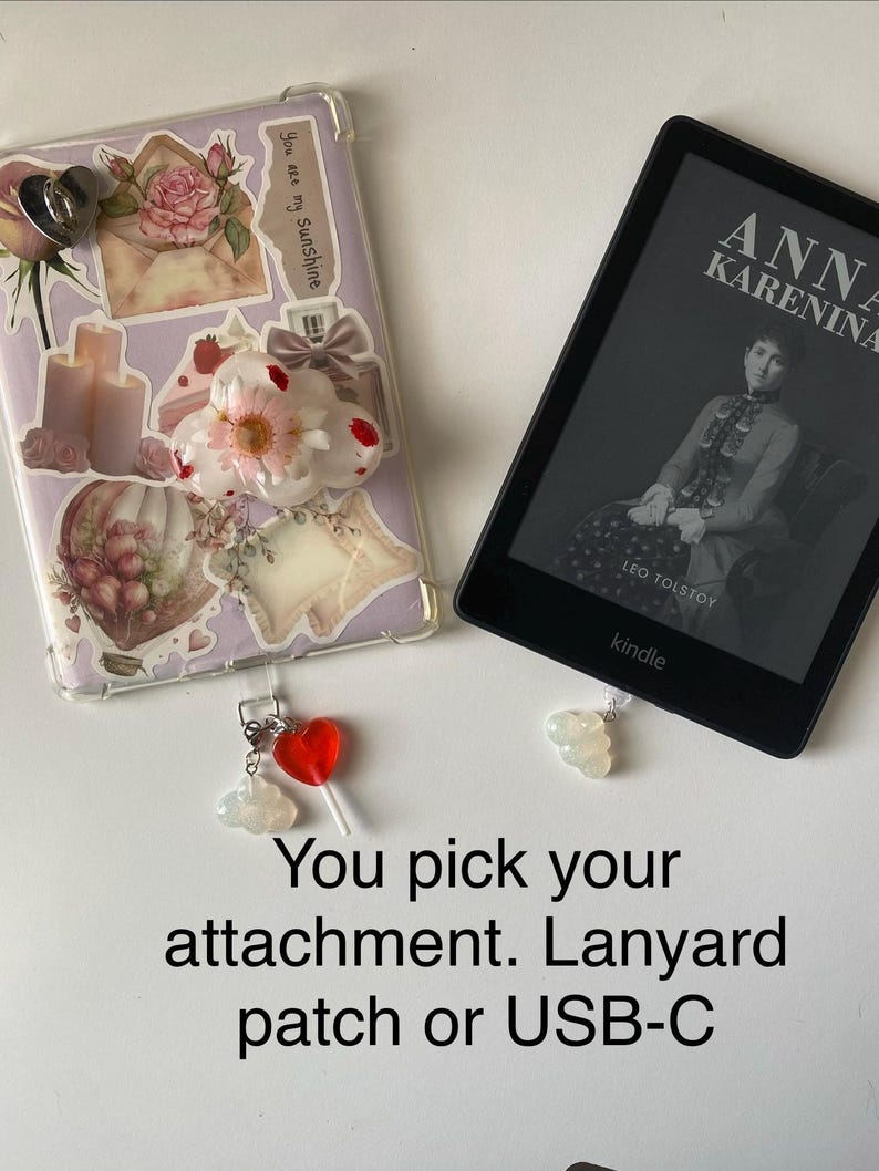 May include: A clear phone case decorated with stickers, including a rose, envelope, and candles. A Kindle e-reader displays the cover of "Anna Karenina." Charms include a red heart lollipop and a cloud. Text reads "You pick your attachment. Lanyard patch or USB-C."