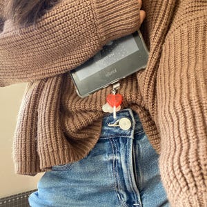 May include: A brown knit sweater and blue denim jeans. A clear phone case holds a Kindle device. A red heart charm hangs from the phone. The Kindle screen displays text.