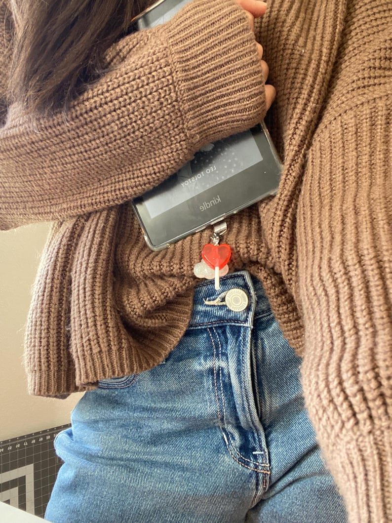 May include: A brown knit sweater and blue denim jeans. A clear phone case holds a Kindle device. A red heart charm hangs from the phone. The Kindle screen displays text.