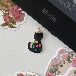 May include: A black cat bookmark with a gold chain and flowers on its back. The bookmark is attached to a black Kindle e-reader.