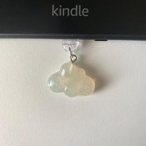 May include: A cloud-shaped charm with glitter, hanging below a Kindle device. The charm is a light, pearlescent color and has a small metal loop for attachment. The word "kindle" is printed on the device.