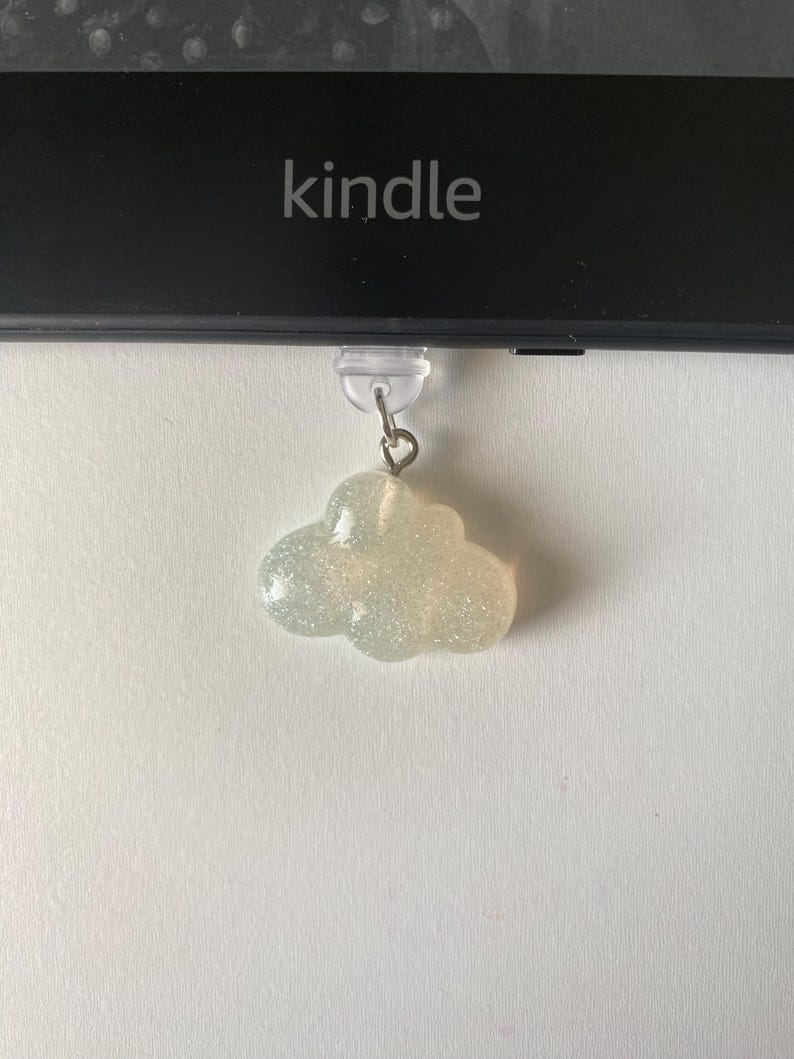 May include: A cloud-shaped charm with glitter, hanging below a Kindle device. The charm is a light, pearlescent color and has a small metal loop for attachment. The word "kindle" is printed on the device.