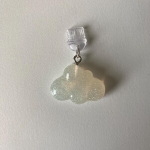 May include: A cloud-shaped earring with a clear post and a silver-colored metal loop. The cloud is a light, iridescent color with glitter embedded throughout. The earring is a whimsical accessory.