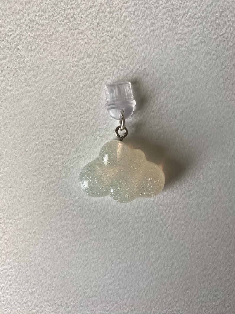 May include: A cloud-shaped earring with a clear post and a silver-colored metal loop. The cloud is a light, iridescent color with glitter embedded throughout. The earring is a whimsical accessory.