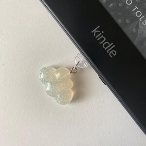May include: A sparkly, cloud-shaped charm attached to a Kindle e-reader. The charm is a light, iridescent color and hangs from a clear plastic clip. The Kindle is black with the word "kindle" printed on it.
