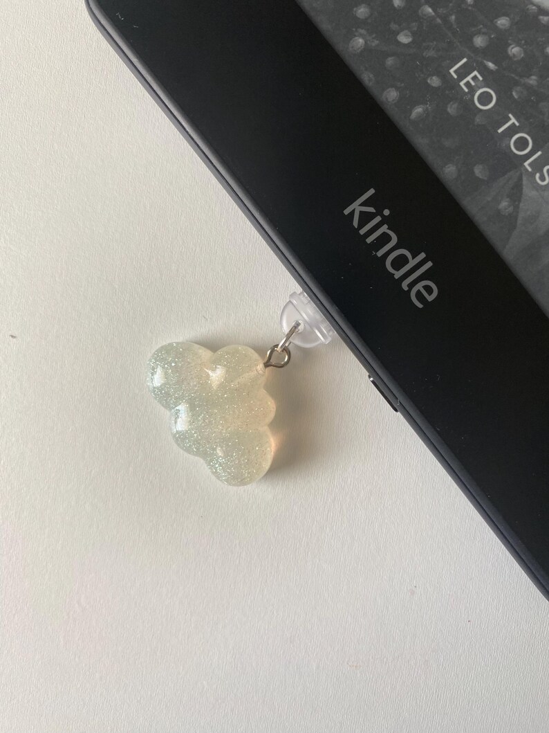 May include: A sparkly, cloud-shaped charm attached to a Kindle e-reader. The charm is a light, iridescent color and hangs from a clear plastic clip. The Kindle is black with the word "kindle" printed on it.