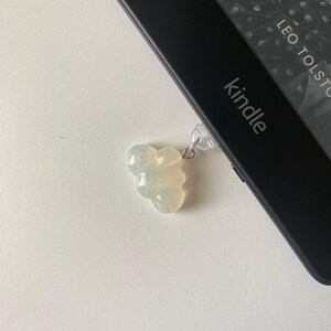 May include: A cloud-shaped, glittery, light-colored charm attached to a Kindle e-reader. The e-reader is black, and the screen displays text from a book. The word "Kindle" is visible on the device.