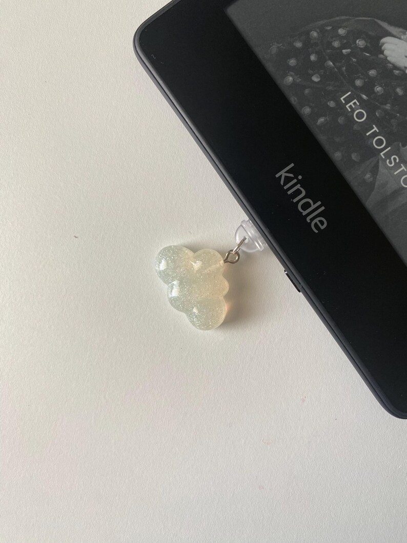 May include: A cloud-shaped, glittery, light-colored charm attached to a Kindle e-reader. The e-reader is black, and the screen displays text from a book. The word "Kindle" is visible on the device.