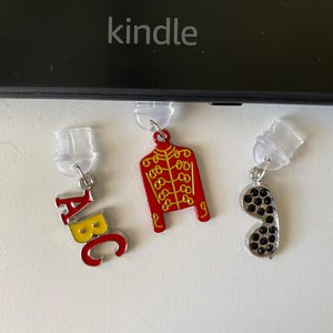 May include: Three metal charms with clear plastic attachments. One charm features the letters ABC in red and yellow. The second charm is a red military-style jacket with gold detailing. The third charm is a silver-toned shape with black dots.