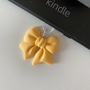 May include: A light yellow bow charm with a silver-colored clasp. The bow is made of a smooth material and is attached to a black Kindle device. The word "kindle" is printed on the device.