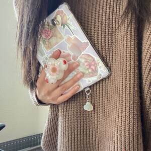 May include: A lavender phone case adorned with floral stickers and a white flower phone grip. A cloud charm hangs from the case. The person is wearing a brown sweater.