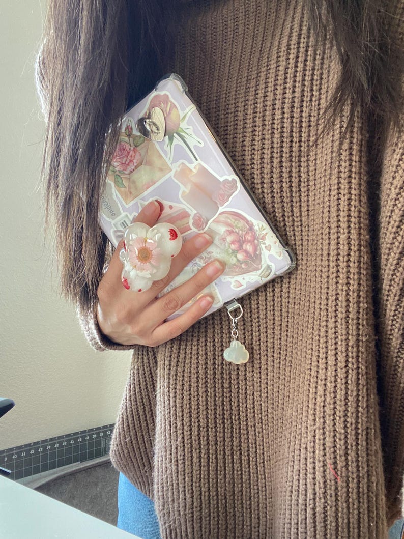 May include: A lavender phone case adorned with floral stickers and a white flower phone grip. A cloud charm hangs from the case. The person is wearing a brown sweater.