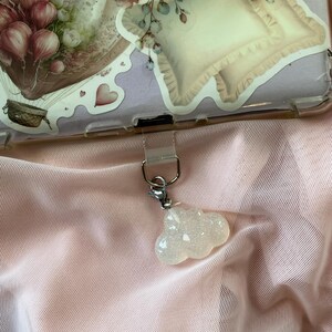 May include: A clear phone case with floral stickers, including a hot air balloon and pillow design. A small, glittery cloud charm hangs from the case. The background is a sheer, light pink fabric.