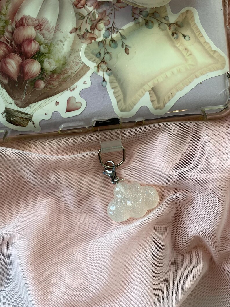 May include: A clear phone case with floral stickers, including a hot air balloon and pillow design. A small, glittery cloud charm hangs from the case. The background is a sheer, light pink fabric.