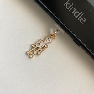 May include: A gold-colored skeleton charm with white enamel detailing hangs from a clear plastic attachment. The charm is positioned near a black Kindle device, with the word "kindle" printed on the device.