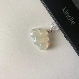 May include: A glittery, cloud-shaped charm with a silver-colored metal loop attached to a black Kindle device. The Kindle has the word "kindle" printed on it. The charm is a light, iridescent color.