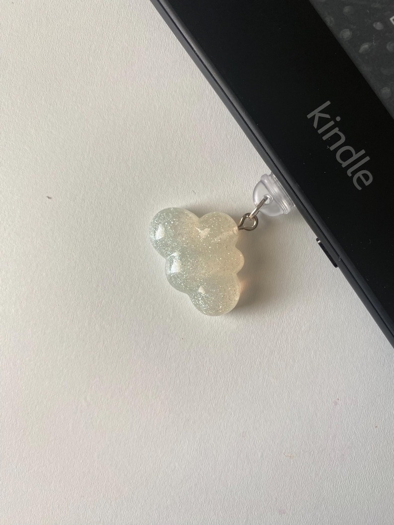May include: A glittery, cloud-shaped charm with a silver-colored metal loop attached to a black Kindle device. The Kindle has the word "kindle" printed on it. The charm is a light, iridescent color.